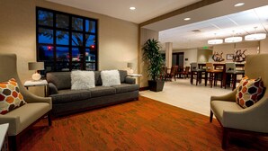 A hotel lobby with a grey sofa, two armchairs, a large window, a dining area, and a potted plant. - Hampton Inn Santa Barbara/Goleta (Goleta)