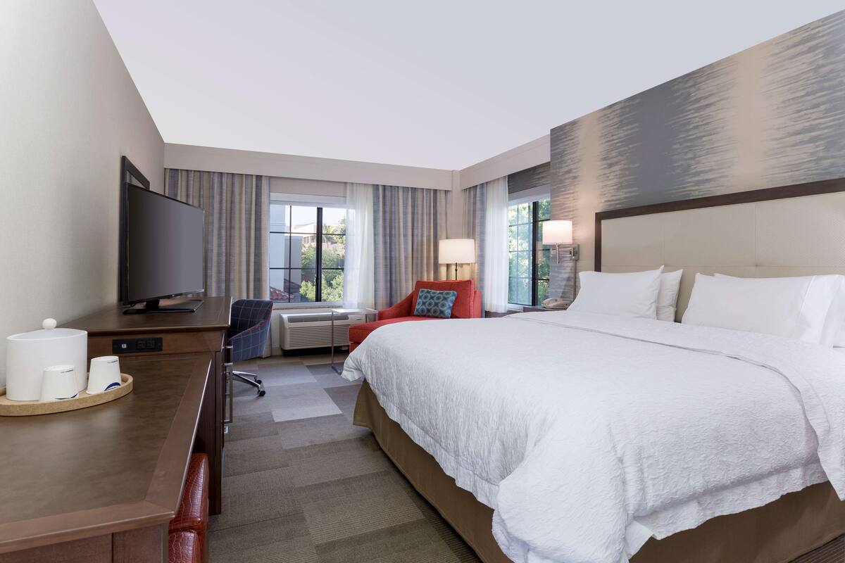 A hotel room with a large bed, a desk with a television, a chair, and a window with curtains.