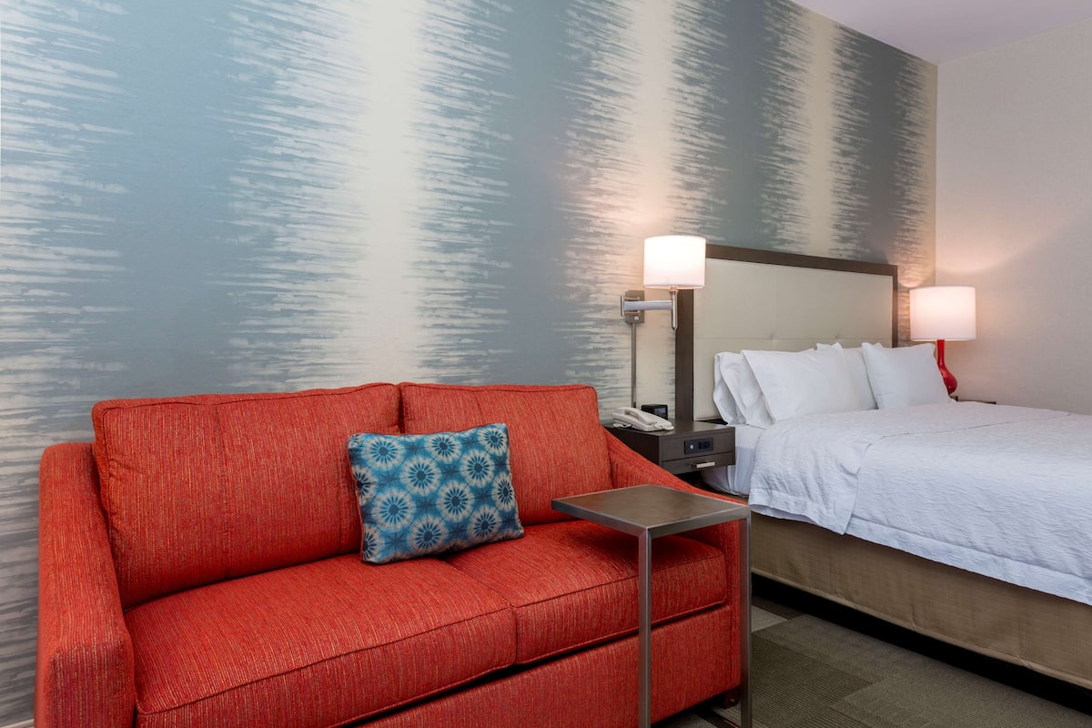 A hotel room with a red sofa, a bed with white bedding, a nightstand with a lamp, and a wall with a textured pattern.