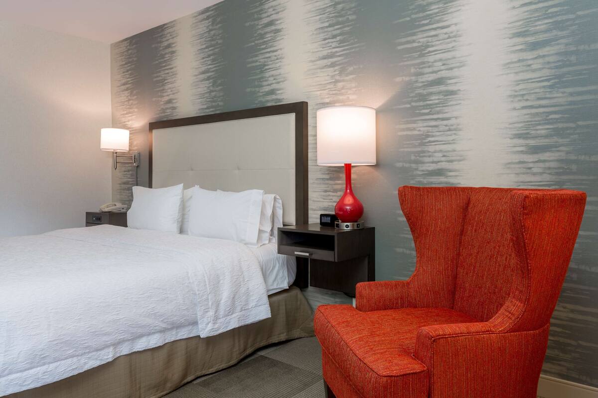 A hotel room with a bed, a nightstand with a lamp, a red chair, and a wall with a textured pattern.
