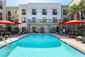 A swimming pool with red umbrellas, lounge chairs, and a building with balconies in the background.