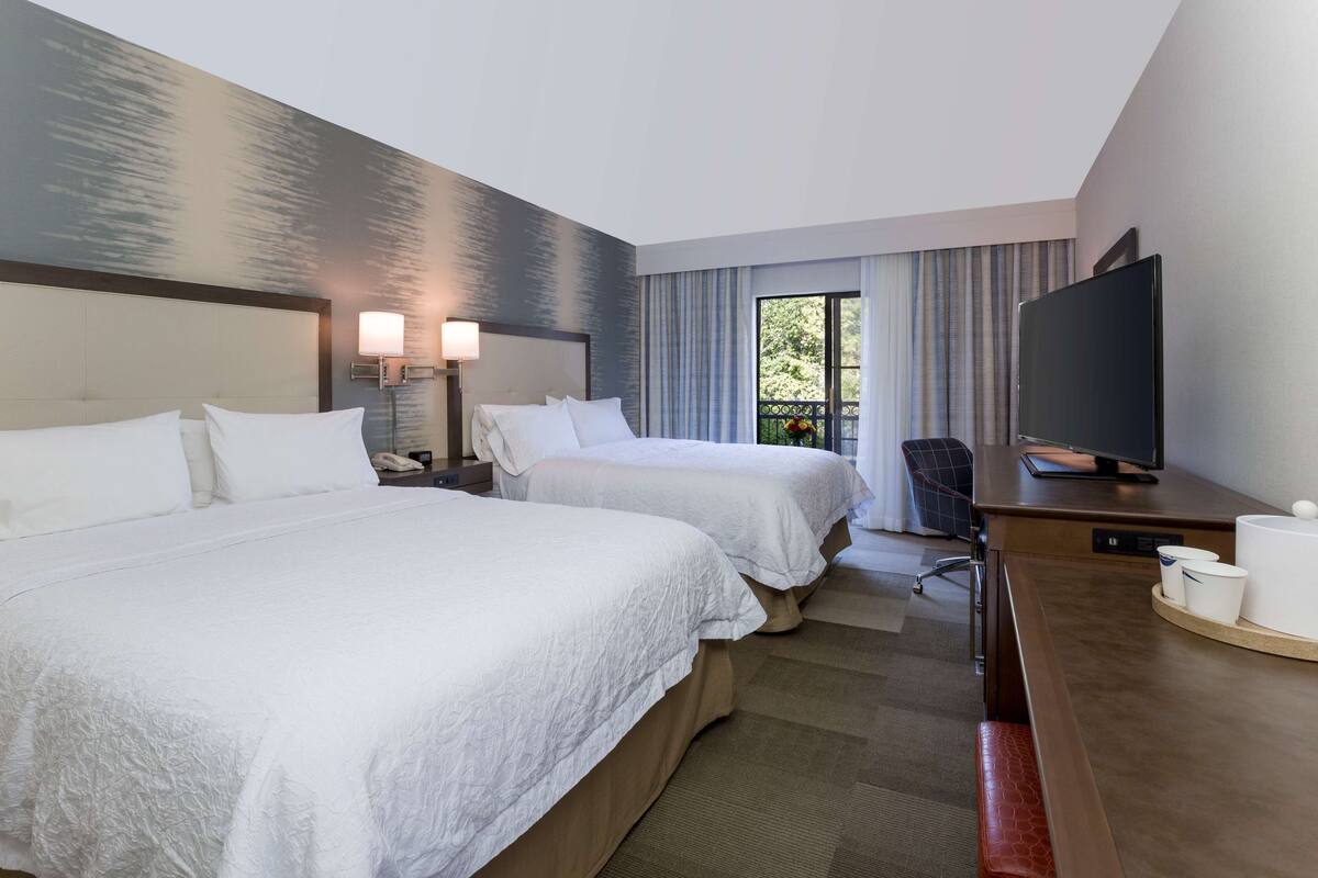 A hotel room with two beds, a desk with a TV, a lamp, and a window with curtains.