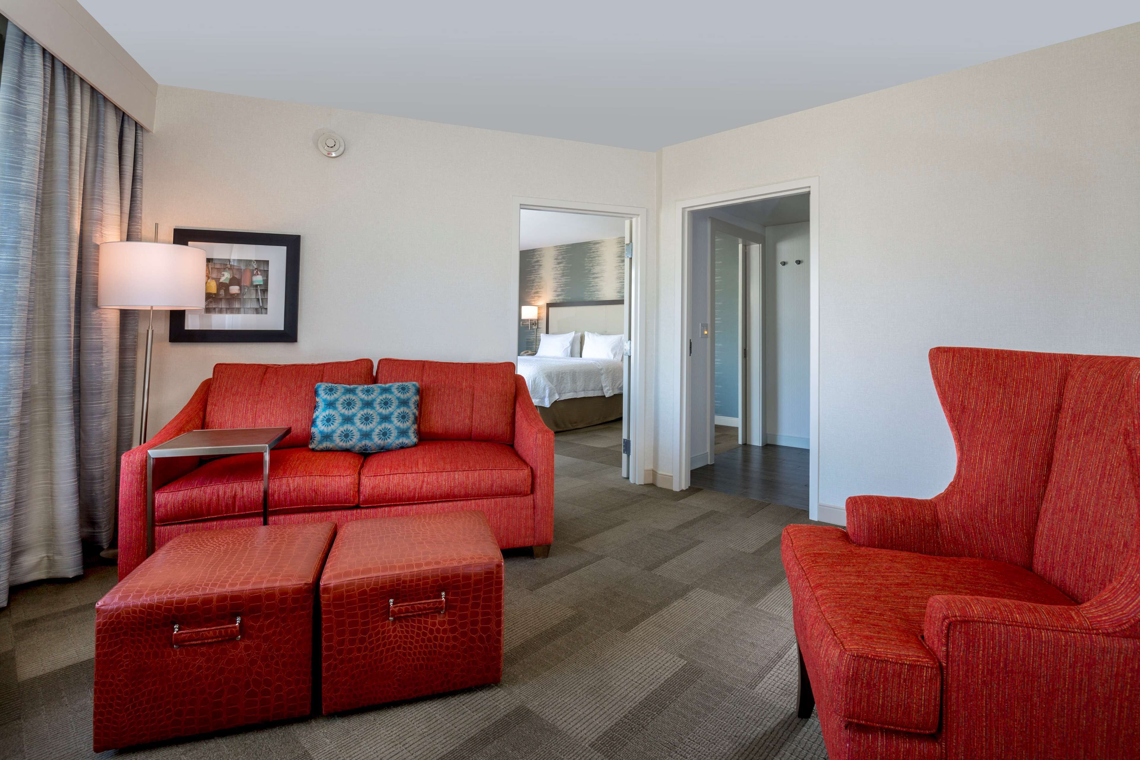 A hotel room with a red sofa, a matching ottoman, a small table, and a bed in the background.