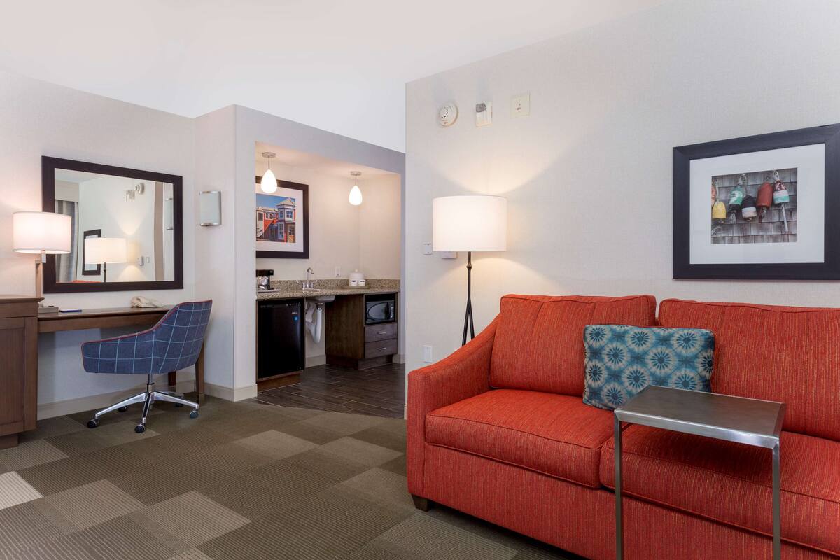 A hotel room with a red sofa, a desk with a chair, a small table, a mirror, a lamp, and a framed picture on the wall.