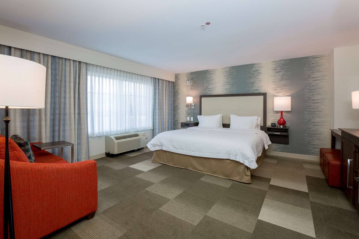 A hotel room with a large bed, a desk, a chair, a lamp, and a heater.