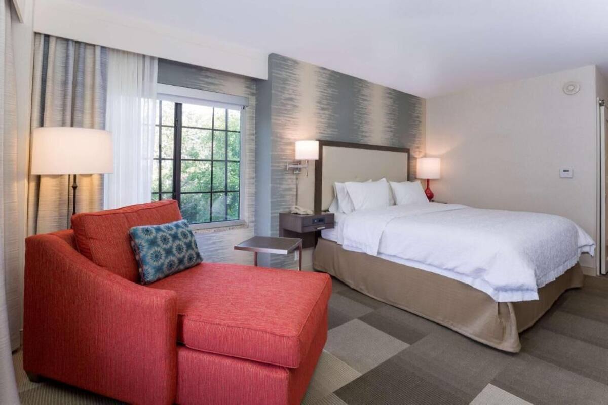 A hotel room with a bed, a red sofa, a nightstand, a lamp, and a window with curtains.