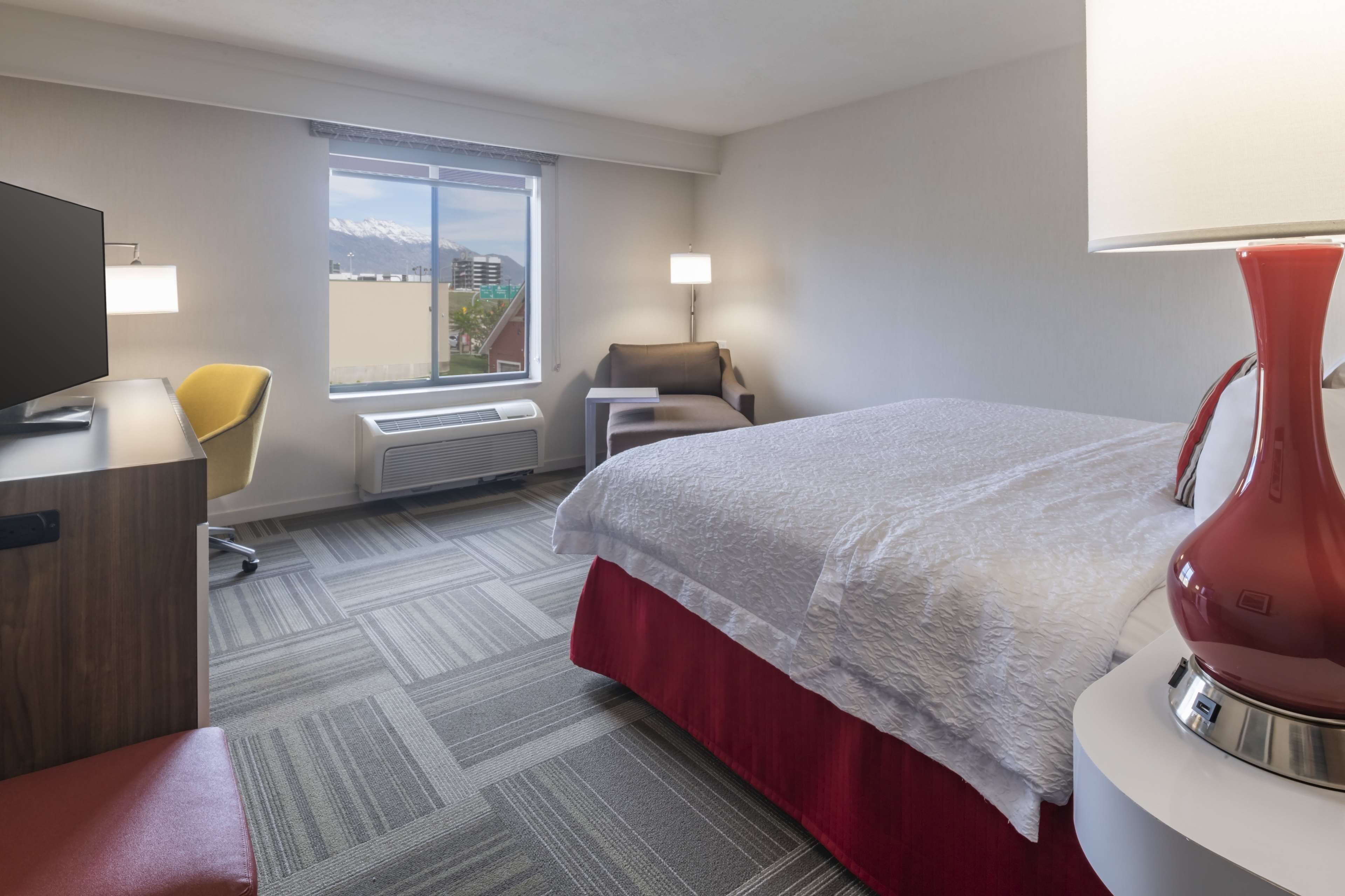 Une chambre d’hôtel avec un lit, un bureau équipé d’un ordinateur, une chaise, une lampe et une fenêtre donnant sur les montagnes.