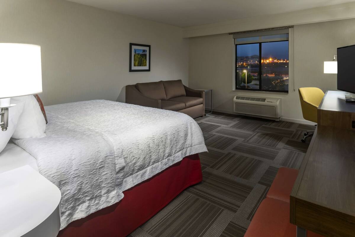 Ein Hotelzimmer mit Bett, Sofa, Schreibtisch und Fenster mit Blick auf die Stadt.