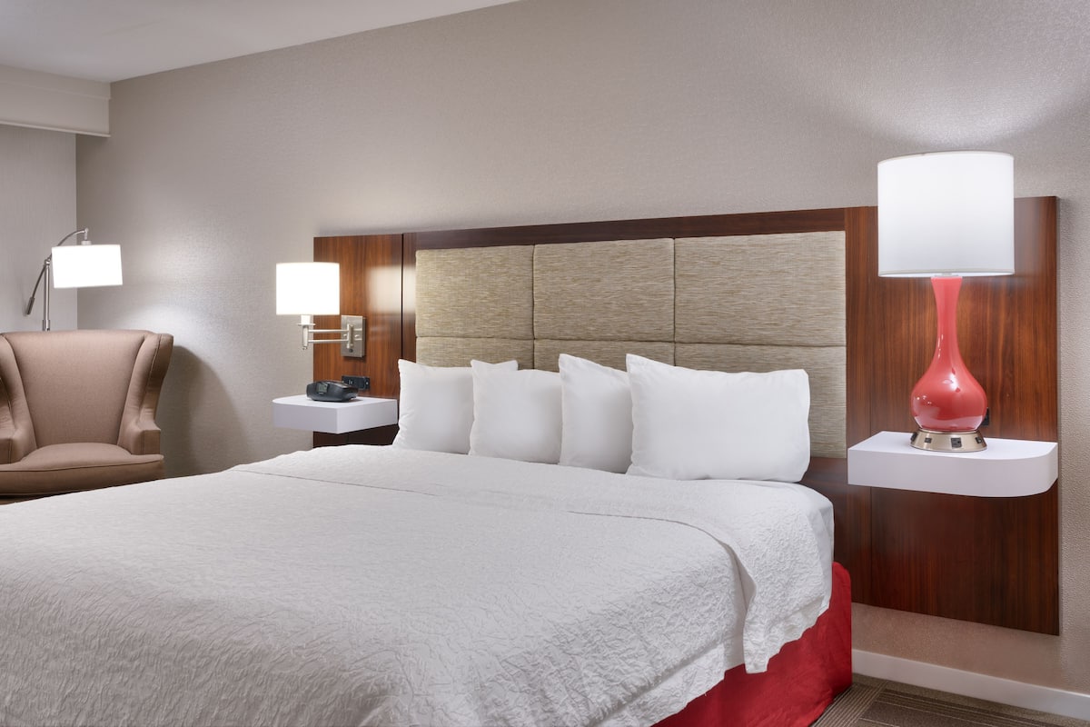 Ein Hotelzimmer mit einem großen Bett, einem Sessel, einem Nachttisch mit Lampe sowie einem Beistelltisch mit Blumenvase.