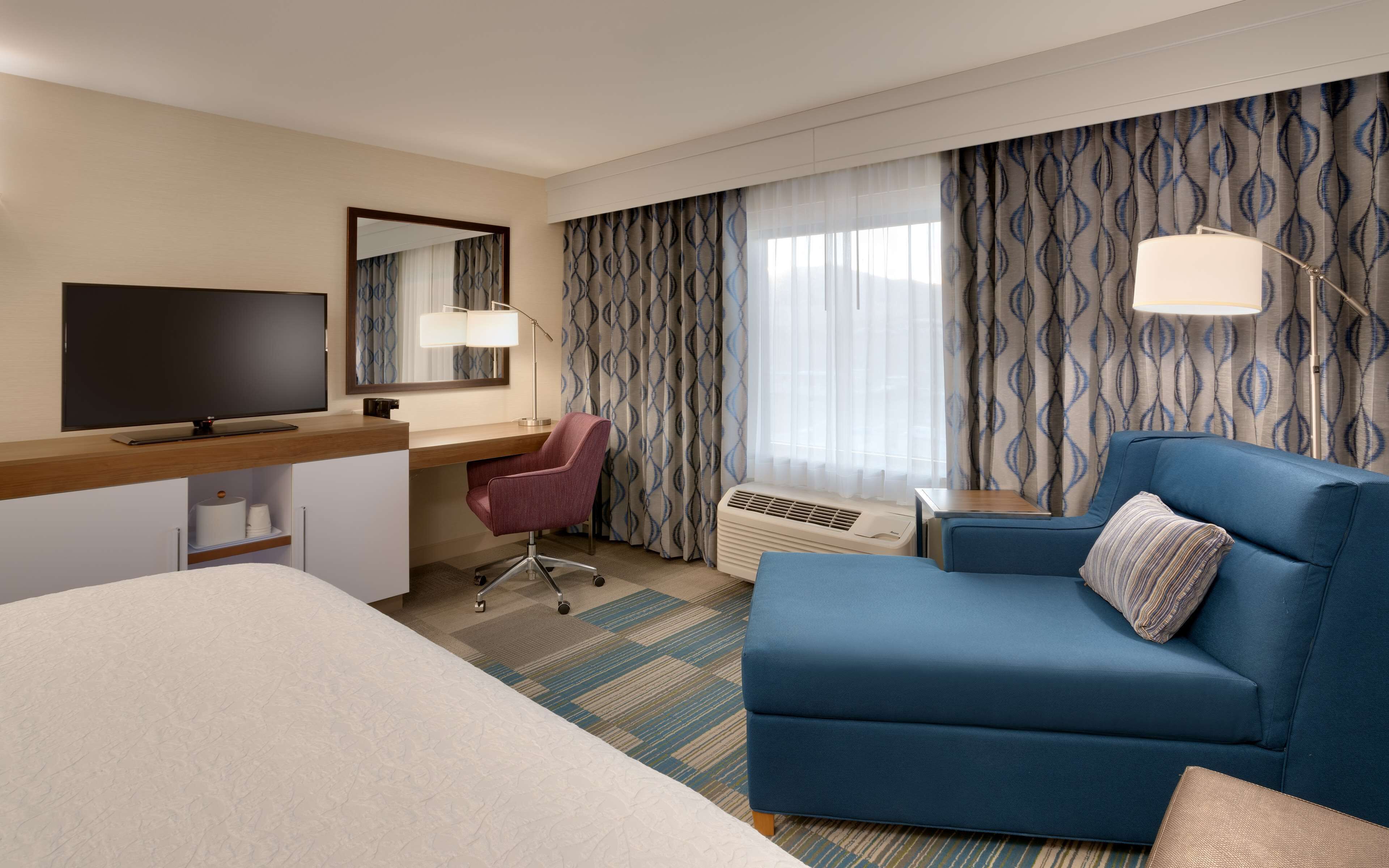 Quarto de hotel com cama, escrivaninha com televisão, sofá azul, cadeira, espelho e cortinas estampadas.