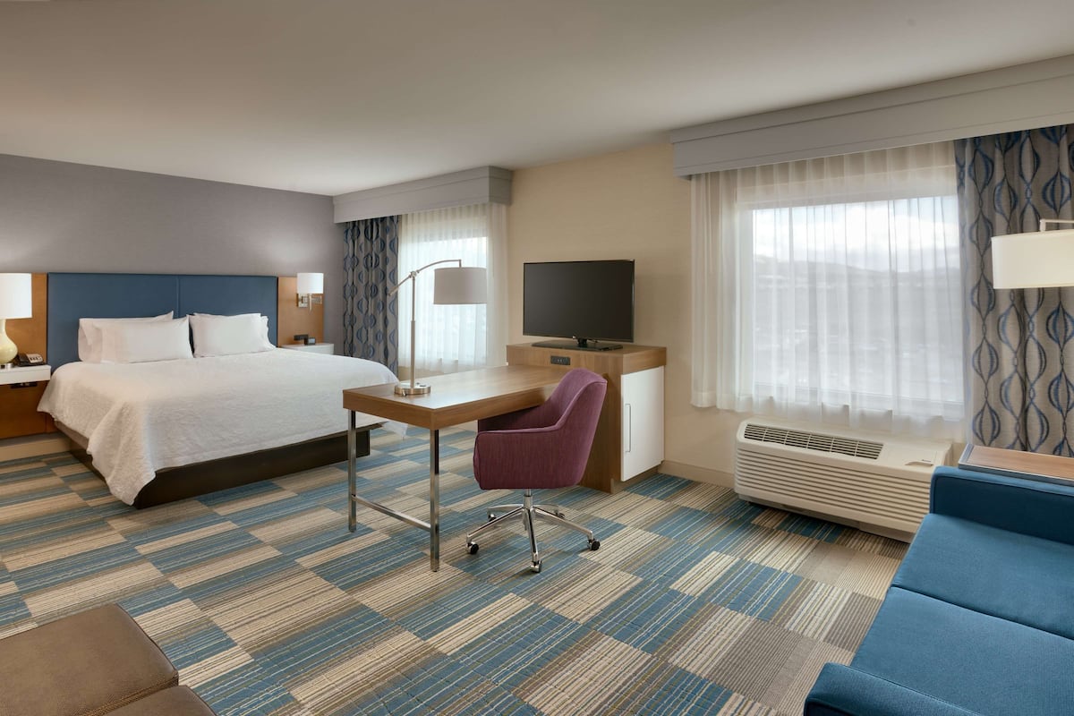 A hotel room with a bed, desk, chair, TV, and air conditioning unit.
