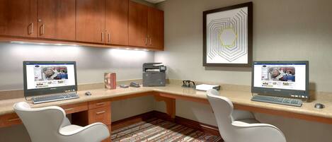 A modern office space with two desks, each with a computer, a printer, and a telephone. There are two white chairs and a patterned carpet.