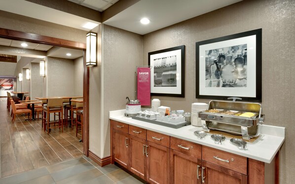 Un espace pour le petit-déjeuner à l’hôtel, avec des tables et des chaises en bois, un buffet proposant divers plats et des tableaux encadrés accrochés au mur.