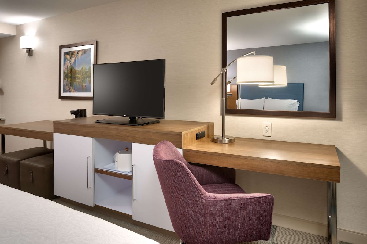 A hotel room with a wooden desk, a flat-screen TV, a purple chair, a desk lamp, a framed picture, and a large mirror.