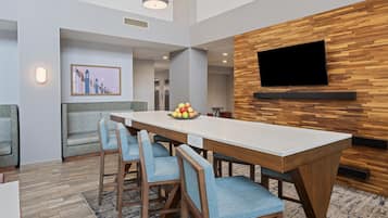 A modern dining area with a long table, blue upholstered chairs, a fruit bowl, and a mounted TV on a wooden wall.