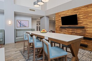 Un espace repas moderne avec une longue table, des chaises à l’assise rembourrée de couleur bleue, un panier de fruits et un téléviseur fixé au mur en bois.