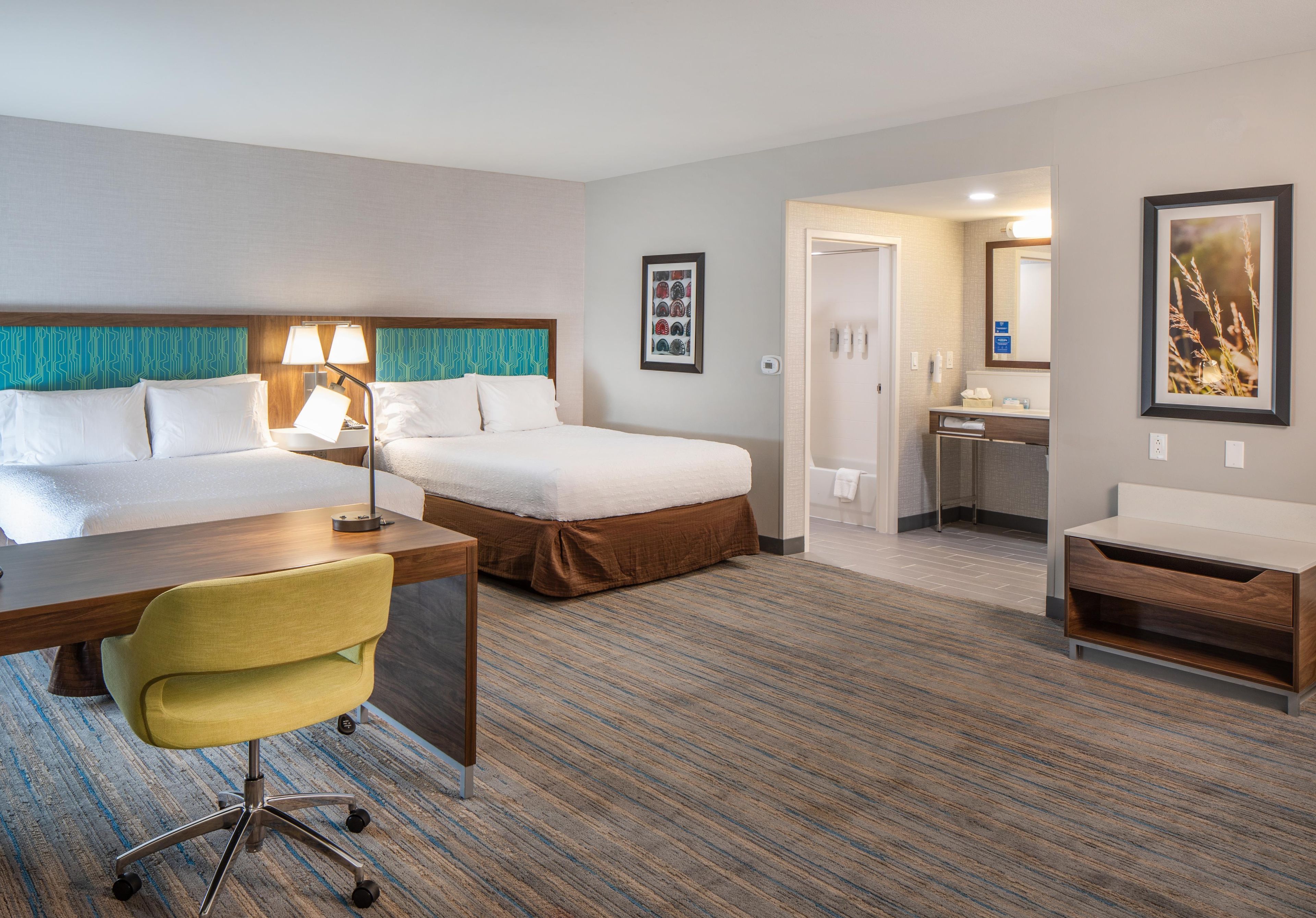 A hotel room with a bed, a desk, a chair, a bathroom, and a framed picture on the wall.