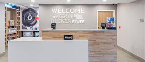 Una mesa de recepción con un cartel que dice 'WELCOME TO THE CORN HUSKER STATE', un terminal de pago digital y una nevera minibar.