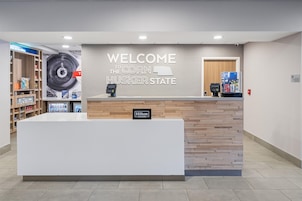 A reception desk with a 'WELCOME TO THE CORN HUSKER STATE' sign, a digital payment terminal, and a minibar.
