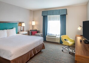 Habitación de hotel con cama, escritorio, silla, lámpara de escritorio, televisor, calefactor y ventana con cortinas.
