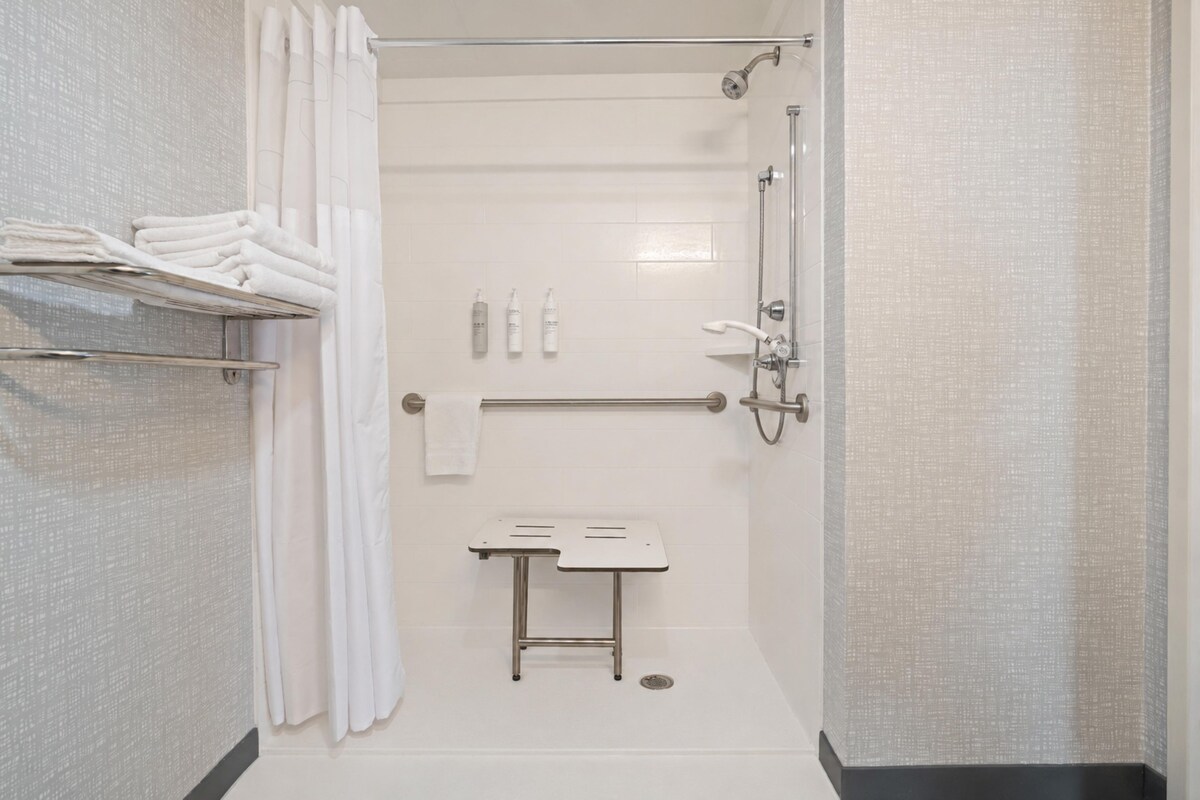 A modern shower area with a glass door, a towel rack with folded towels, a handheld showerhead, and a small table with toiletries.