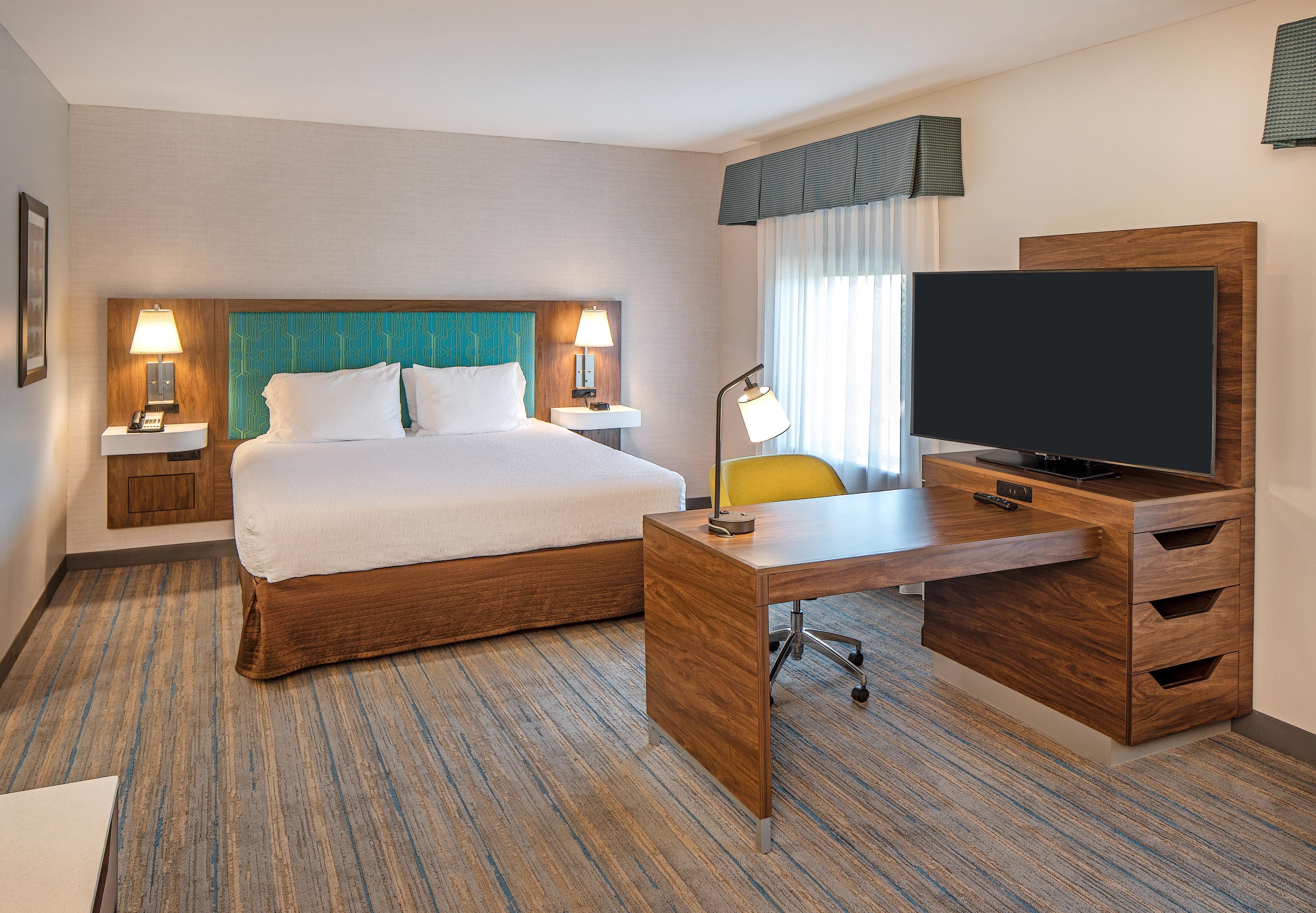Quarto de hotel com cama, escrivaninha com televisão, cadeira e mesa de cabeceira com abajur.