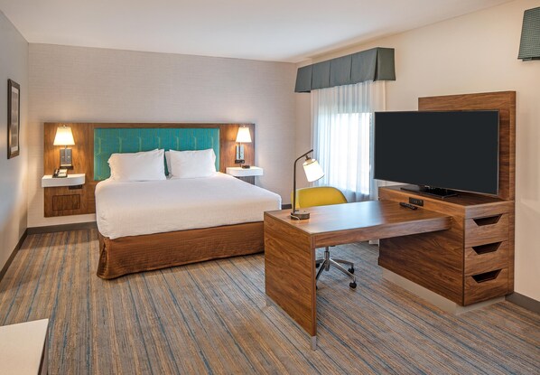 Una habitación de hotel con cama, escritorio con televisor, silla y mesita de noche con lámpara.