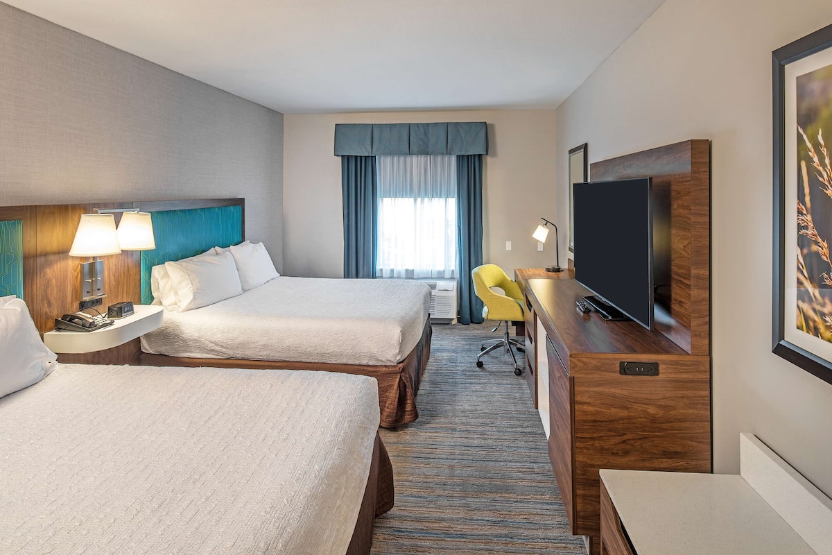 Ett hotellrum med två sängar, ett skrivbord med en tv, en stol och ett stort fönster med gardiner.