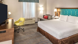 Une chambre d’hôtel avec un grand lit, un bureau avec une chaise, une télévision, une lampe de bureau, une fenêtre avec des rideaux et une lampe fixée au mur.