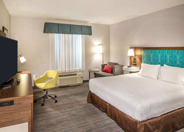 Hotelkamer met een groot bed, een bureau met stoel, een televisie, een bureaulamp, een raam met gordijnen en een wandlamp.