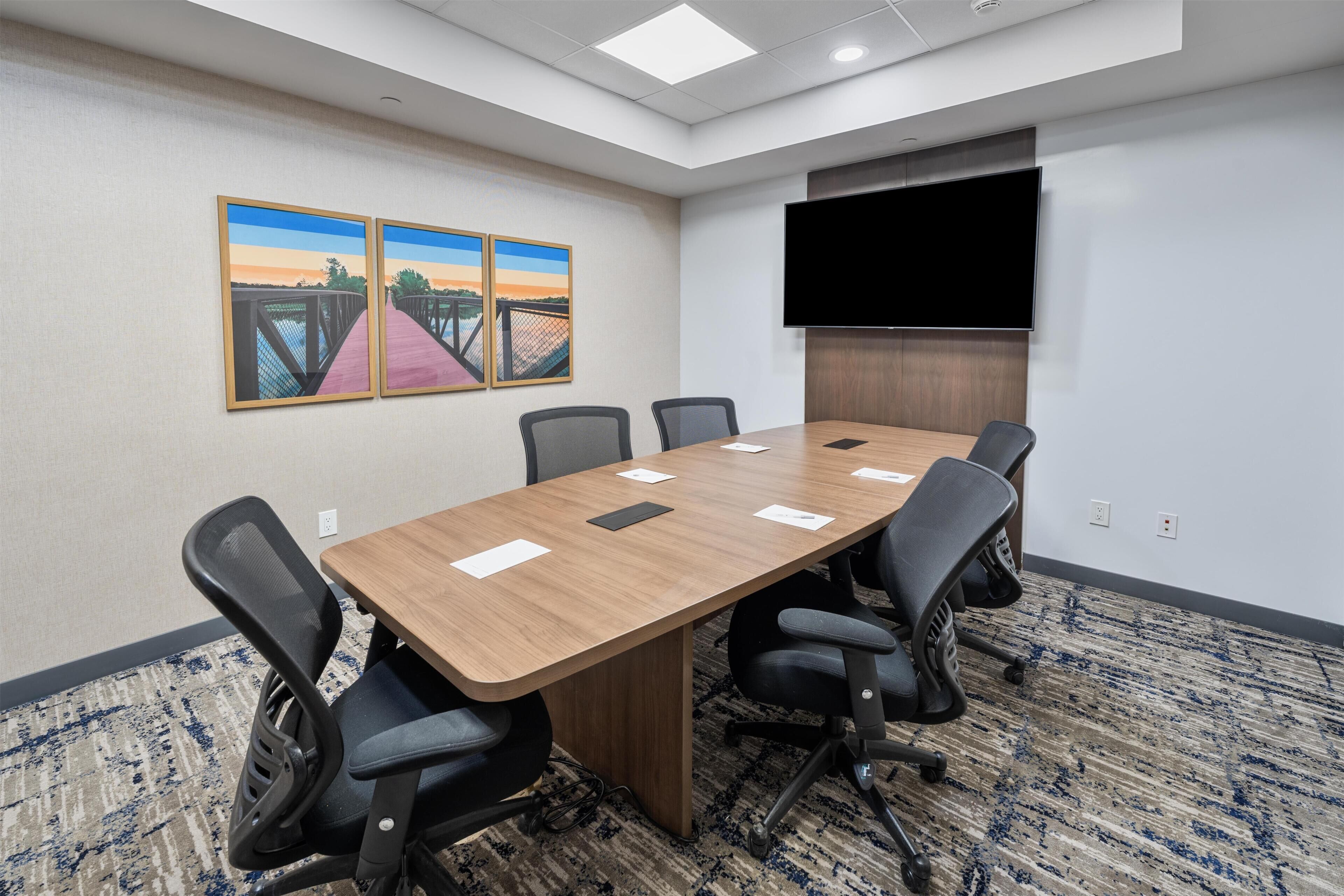 A conference room with a long table, six chairs, a flat-screen TV, and two paintings on the wall.
