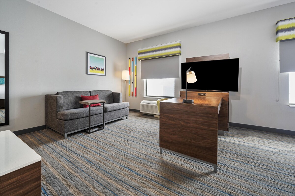 A hotel room with a grey sofa, a wooden desk, a flat-screen TV, a standing lamp, and a window with blinds.