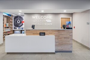 Una mesa de recepción con un cartel que dice 'WELCOME TO THE CORN HUSKER STATE', un terminal de pago digital y una nevera minibar.
