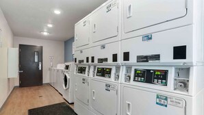 A laundry room with multiple white washing machines and dryers, a door, and a light-colored floor. - Extended Stay America Select Suites - Montgomery (Montgomery)