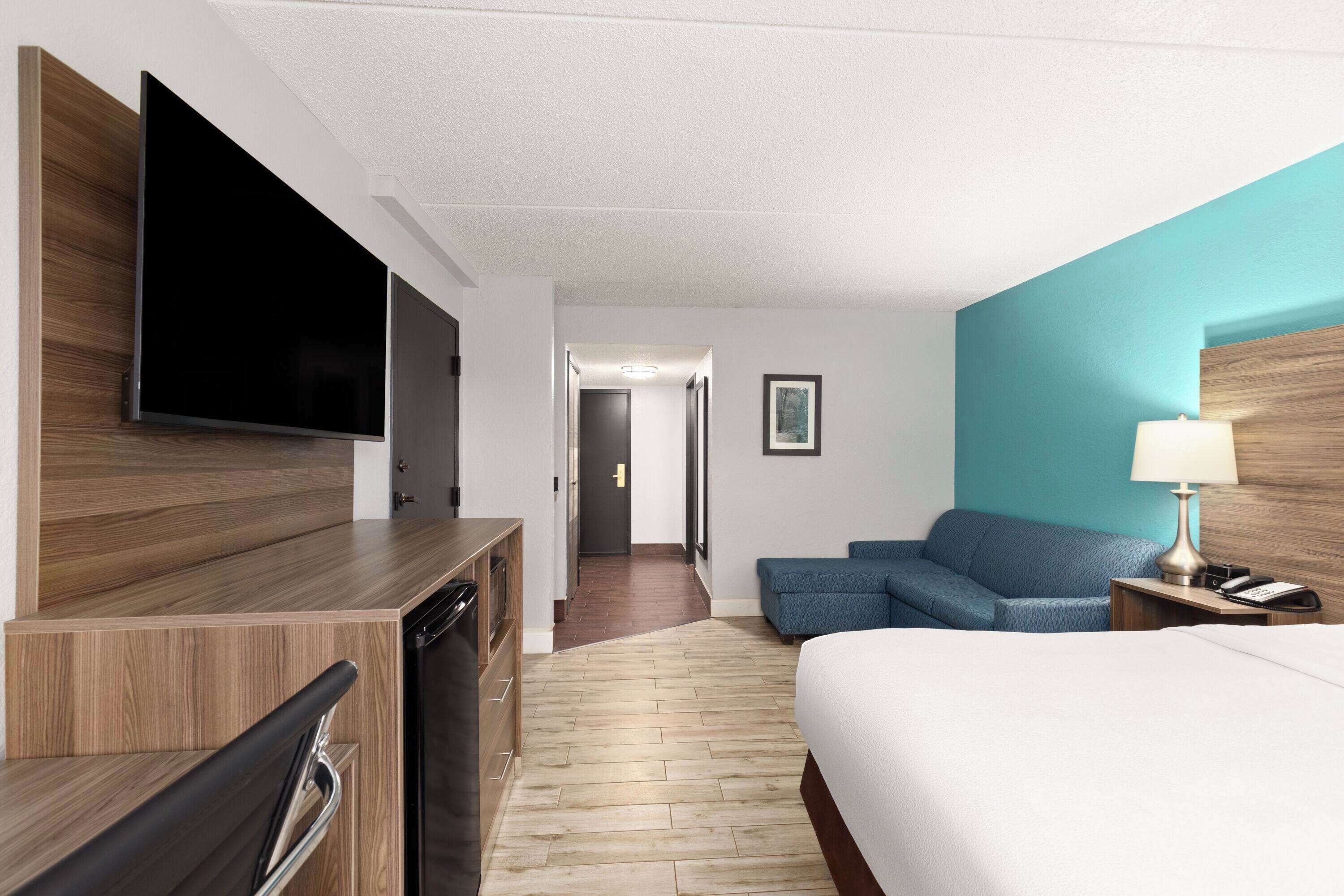 A hotel room with a bed, a TV mounted on the wall, a sofa, a desk with a lamp, and a wooden floor.