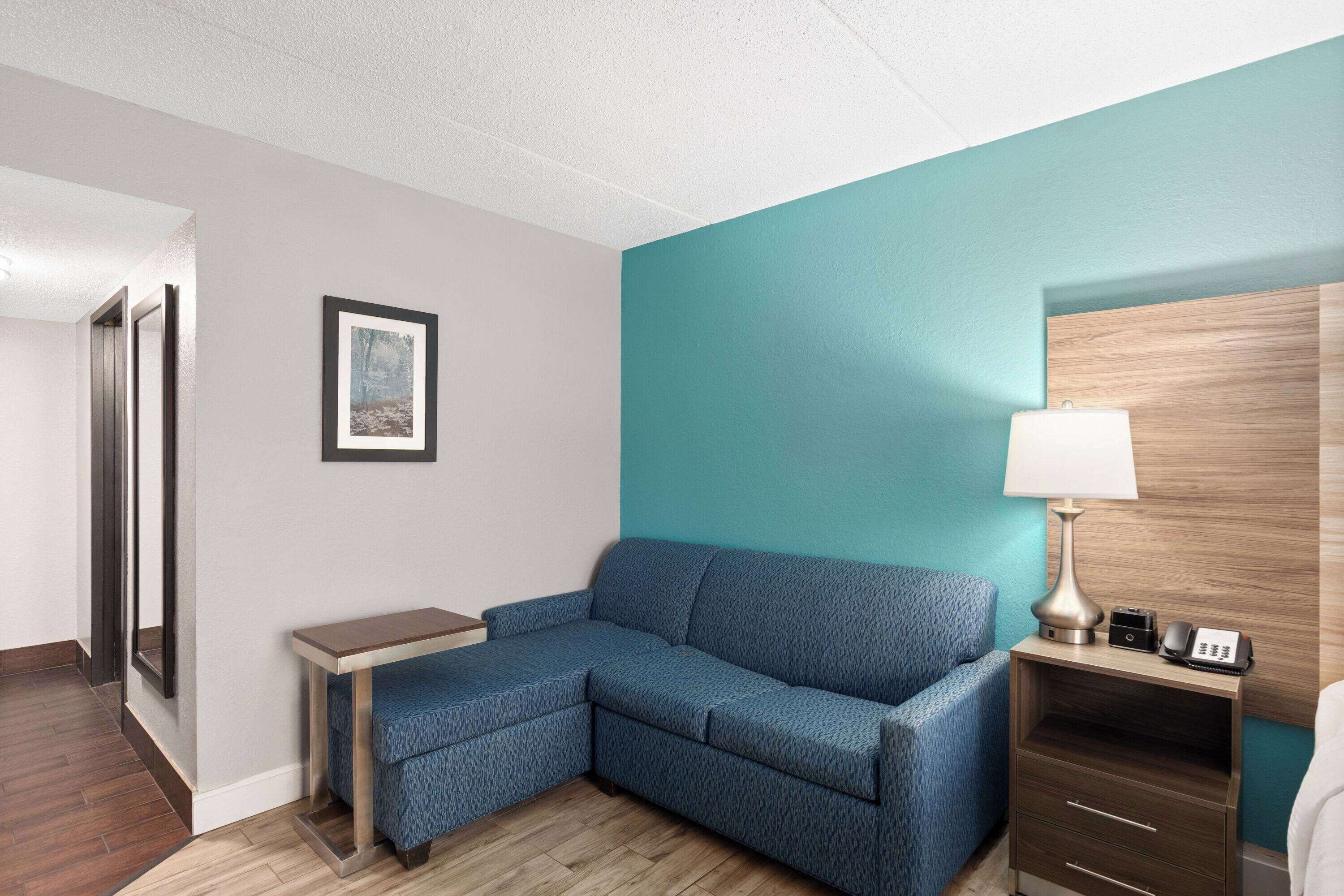A hotel room with a blue sofa, a small table, a lamp, and a wooden nightstand.