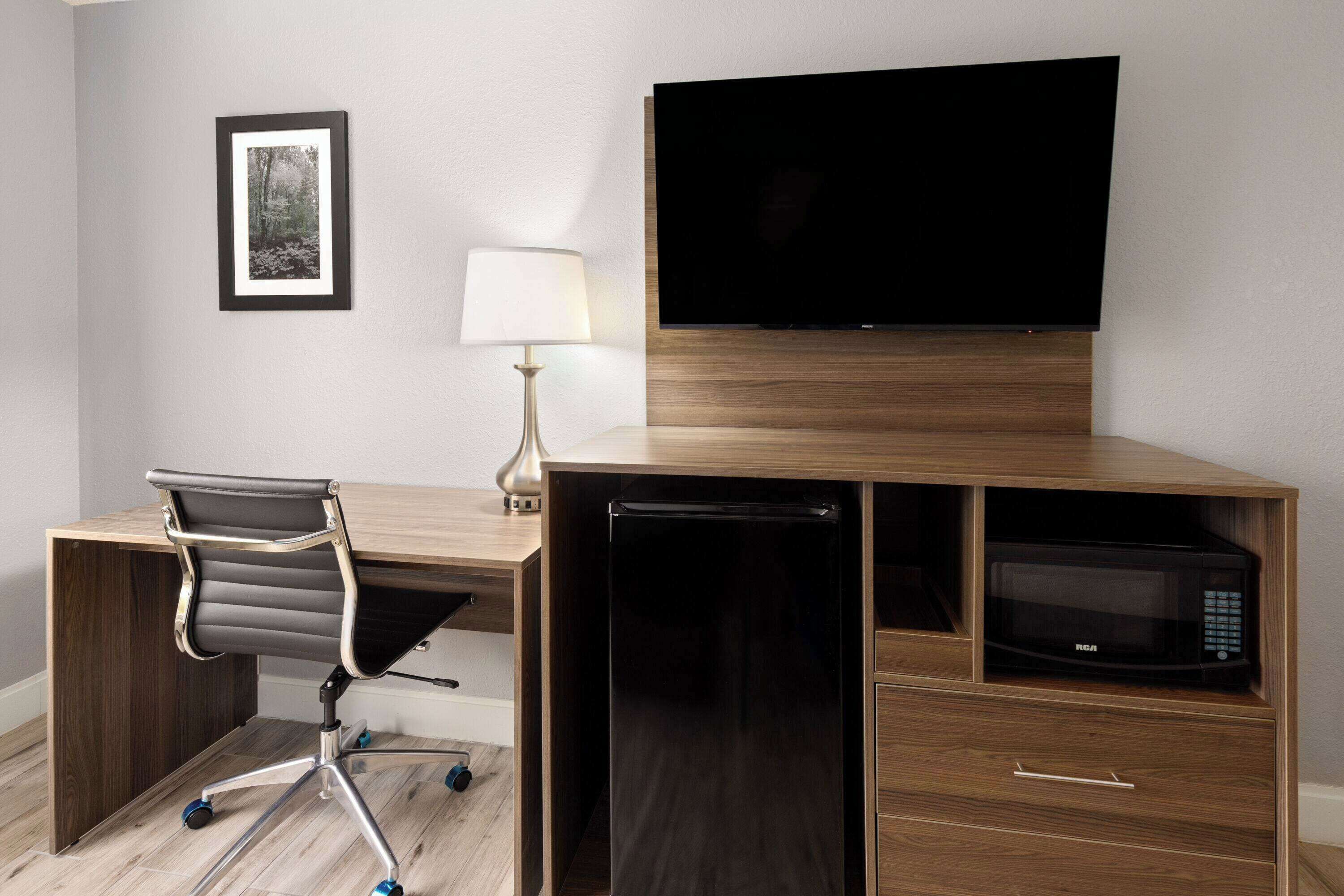 A hotel room with a wooden desk, a black office chair, a flat-screen TV mounted on the wall, a lamp, and a microwave built into the desk.