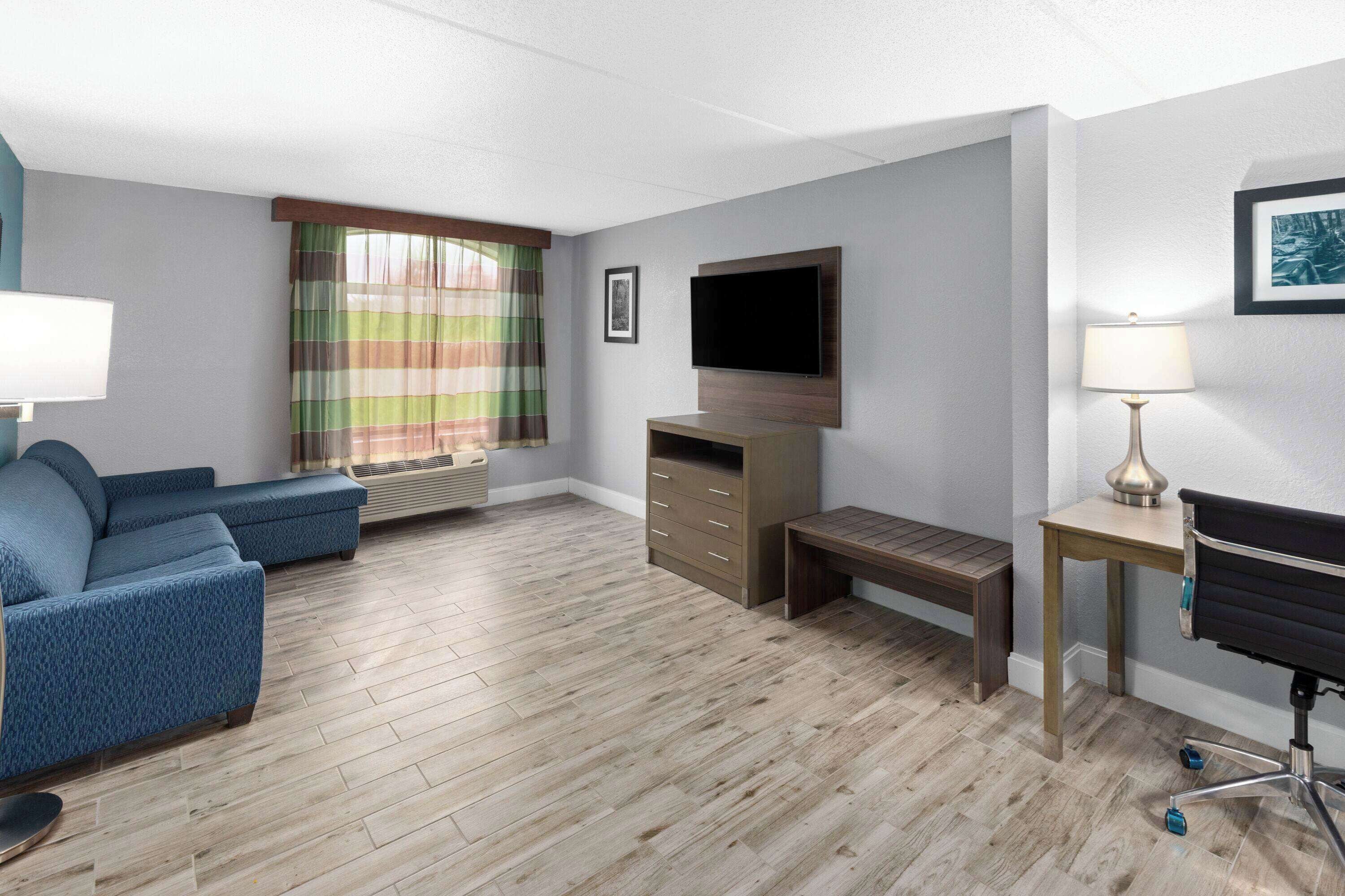 A modern hotel room with a sectional sofa, a desk with a chair, a flat-screen TV mounted on the wall, a small table with a lamp, and a window with striped curtains.