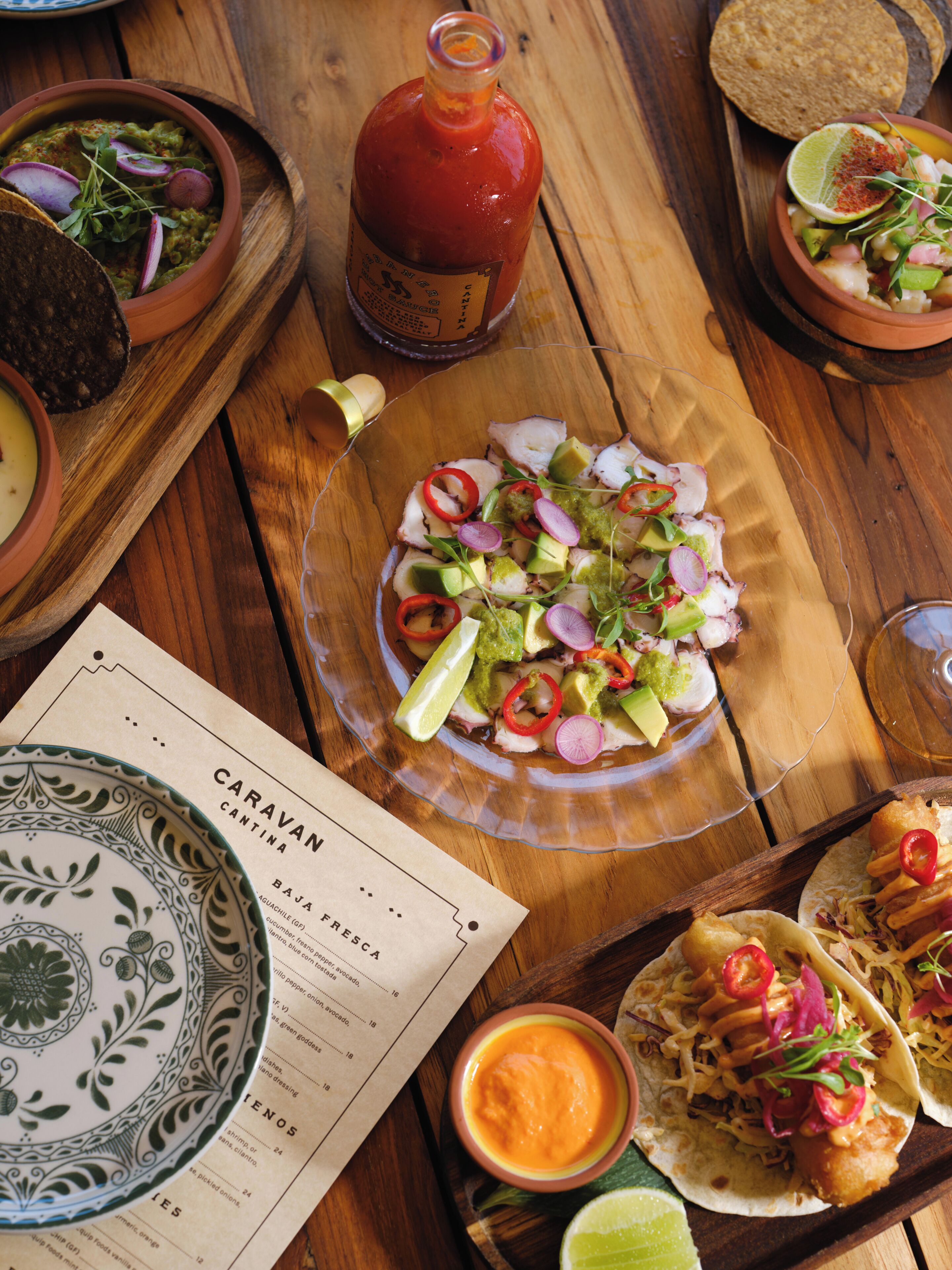 A wooden table with a variety of dishes, including tacos, a salad, and a bottle of sauce.