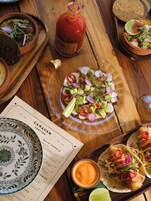 A wooden table with a variety of dishes, including tacos, a salad, and a bottle of sauce.