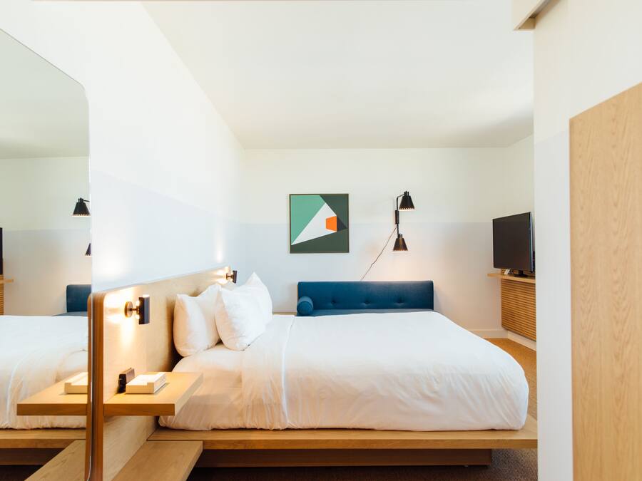 Quarto de hotel moderno com uma cama grande, mesas de cabeceira, televisão e espelho.