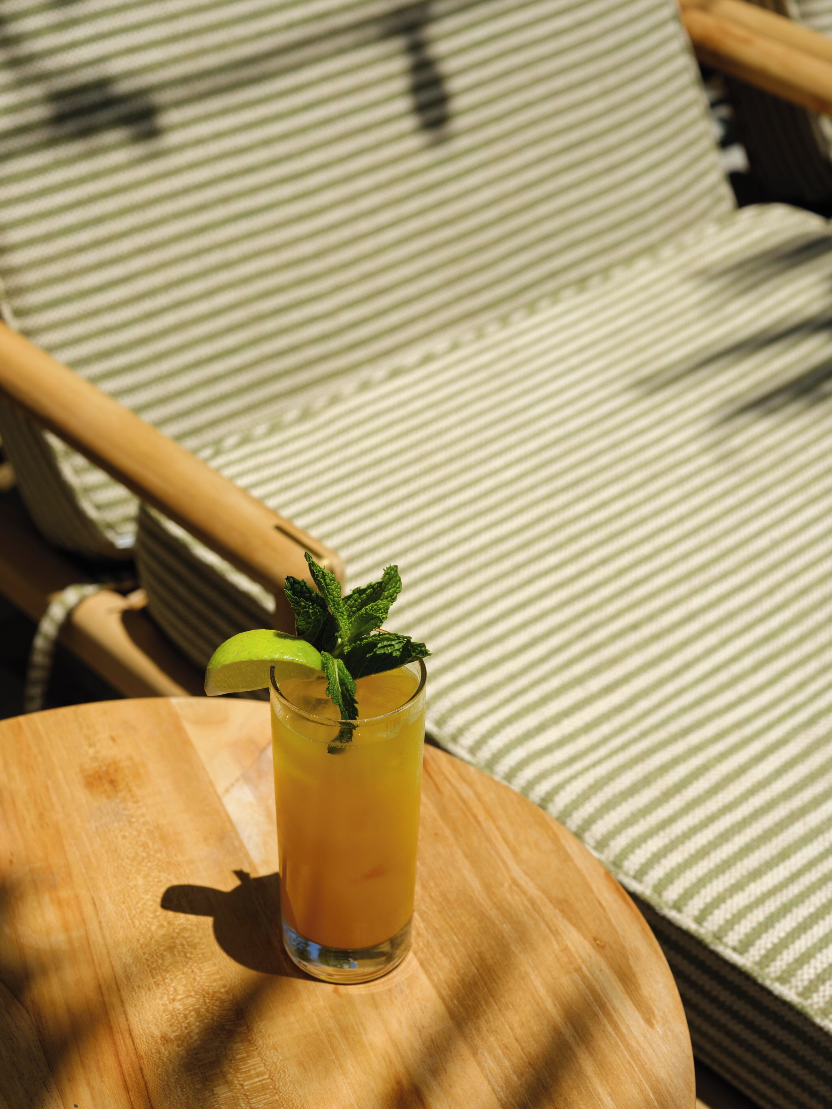 A glass of iced beverage with a lime wedge and mint garnish on a wooden table, set on a striped cushion.