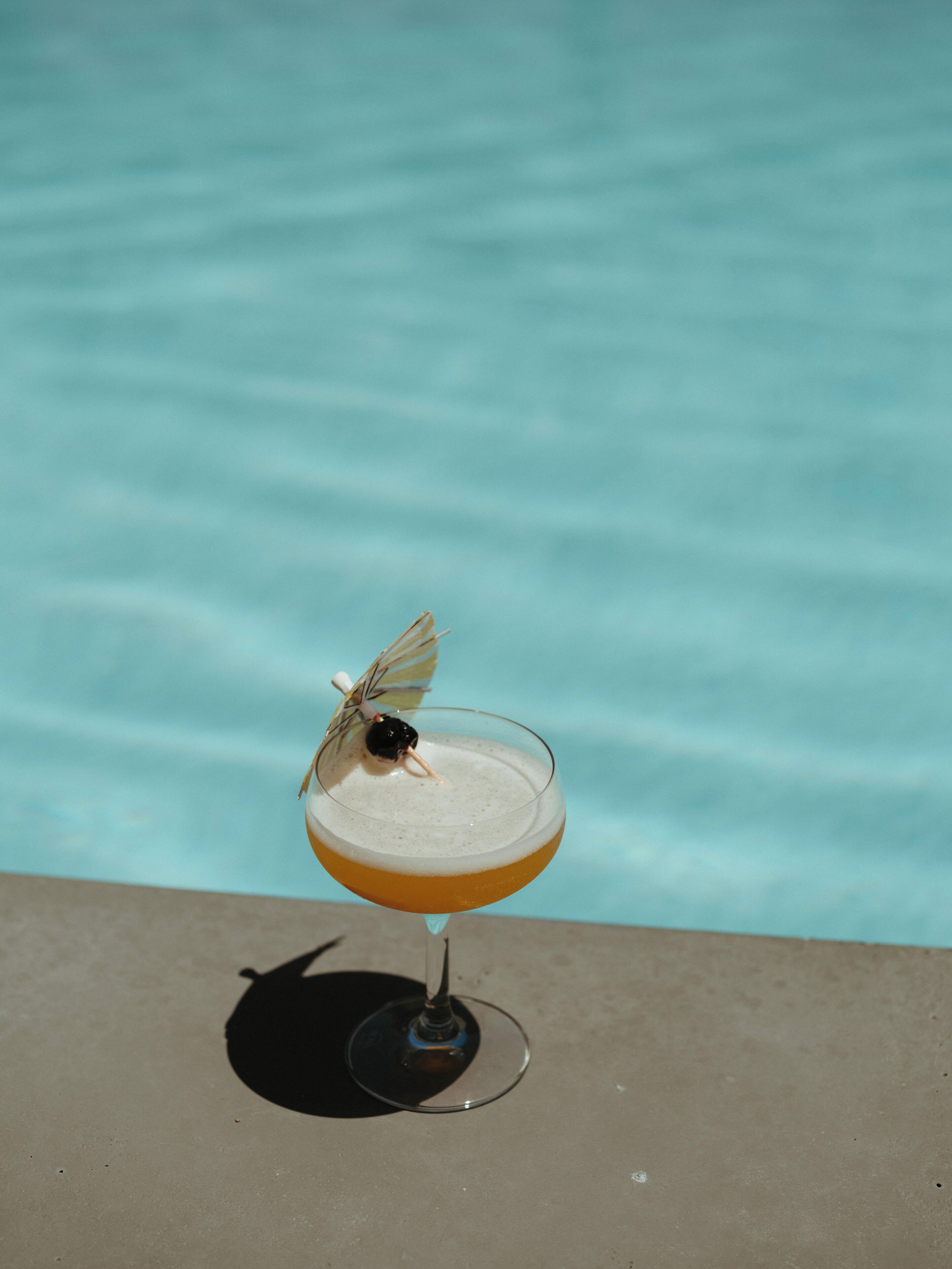 A cocktail glass with a butterfly on the rim, placed on a surface near a pool.