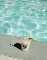 Bebida en vaso con marca, pajita y gajo de lima, sobre superficie de hormigón cerca de una piscina con agua cristalina.