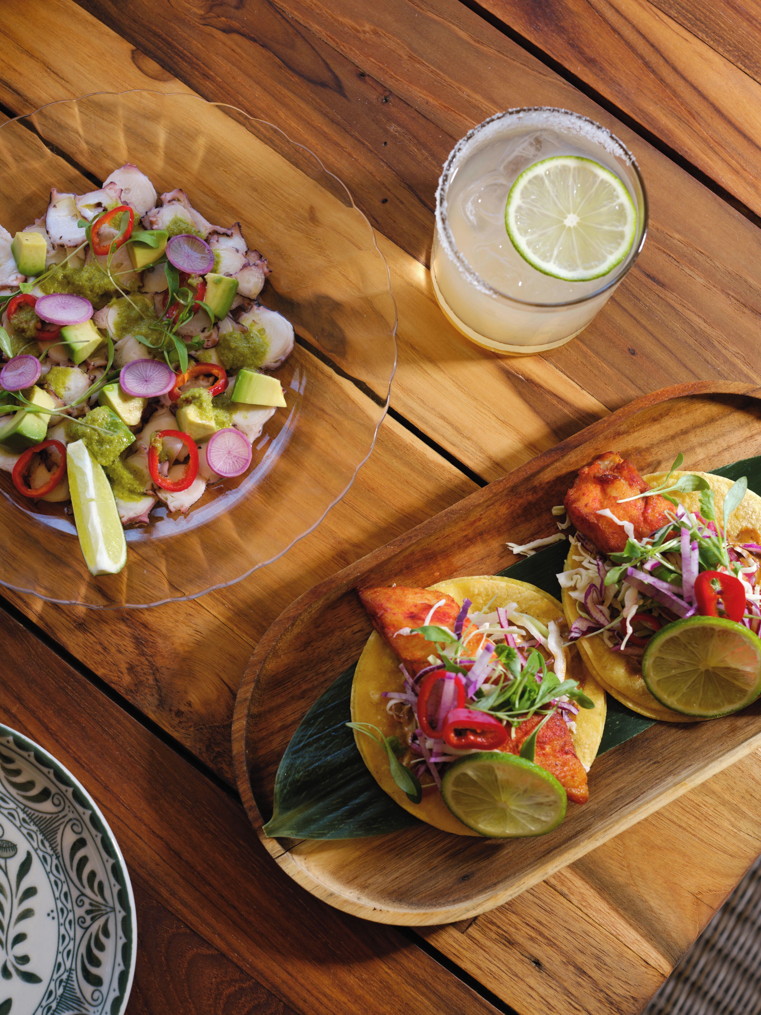 A wooden table with a plate of tacos, a plate of ceviche, and a glass of a lime-based beverage.