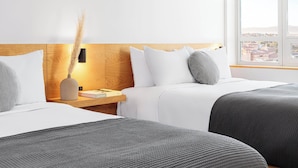 A hotel room with two beds, a nightstand, a vase with dried plants, and a view of a cityscape through a large window. - Hotel June West L.A., a Member of Design Hotels (Los Angeles)