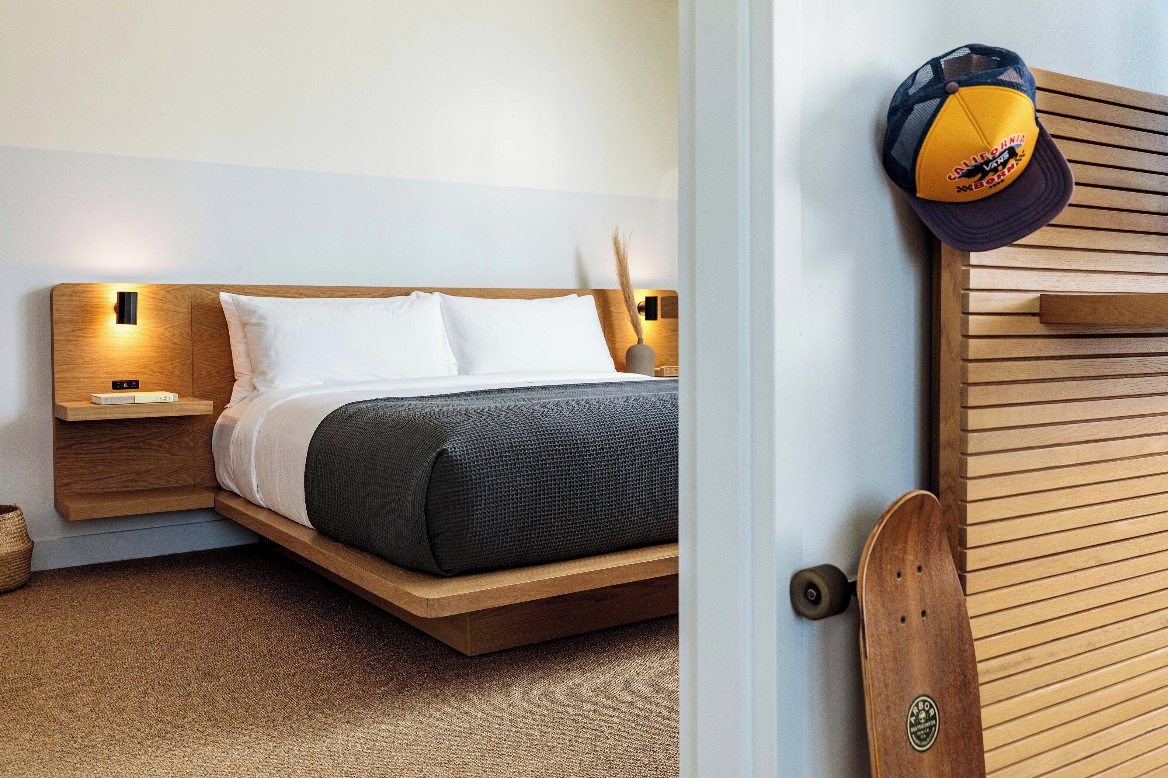 Un dormitorio moderno con cabecera de madera, cama con ropa de cama blanca y oscura, mesita de noche con lámpara, y una puerta de madera con un monopatín apoyado en ella.