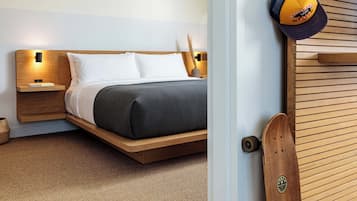 Un dormitorio moderno con cabecera de madera, cama con ropa de cama blanca y oscura, mesita de noche con lámpara, y una puerta de madera con un monopatín apoyado en ella.