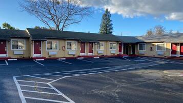 A row of motel buildings with numbered rooms, a parking lot, and a large tree in the background.