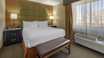 A hotel room with a large bed, bedside tables, a bench, a window with curtains, and an air conditioning unit.